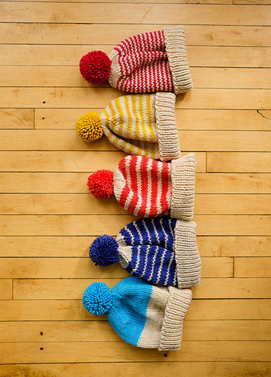 Five knitted Cardo Holiday Hats with pom-poms, featuring red, yellow, and blue stripes or color-block designs, arranged vertically on a wooden floor.