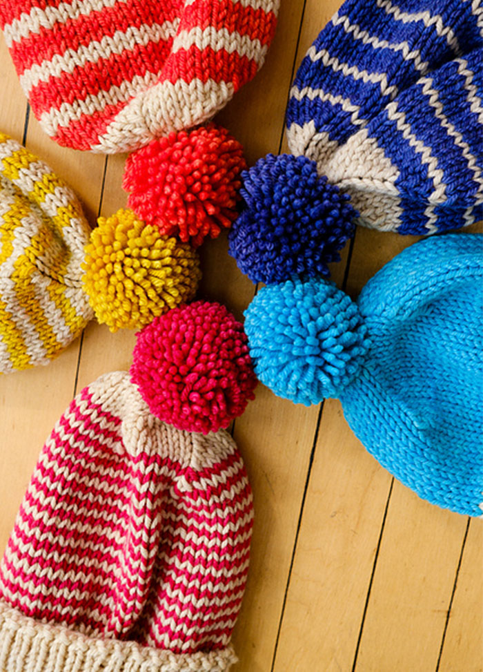 Six knitted Cardo Holiday Hats with pom-poms, in red, blue, yellow, and pink stripes or solids, arranged in a circle on a wooden surface.