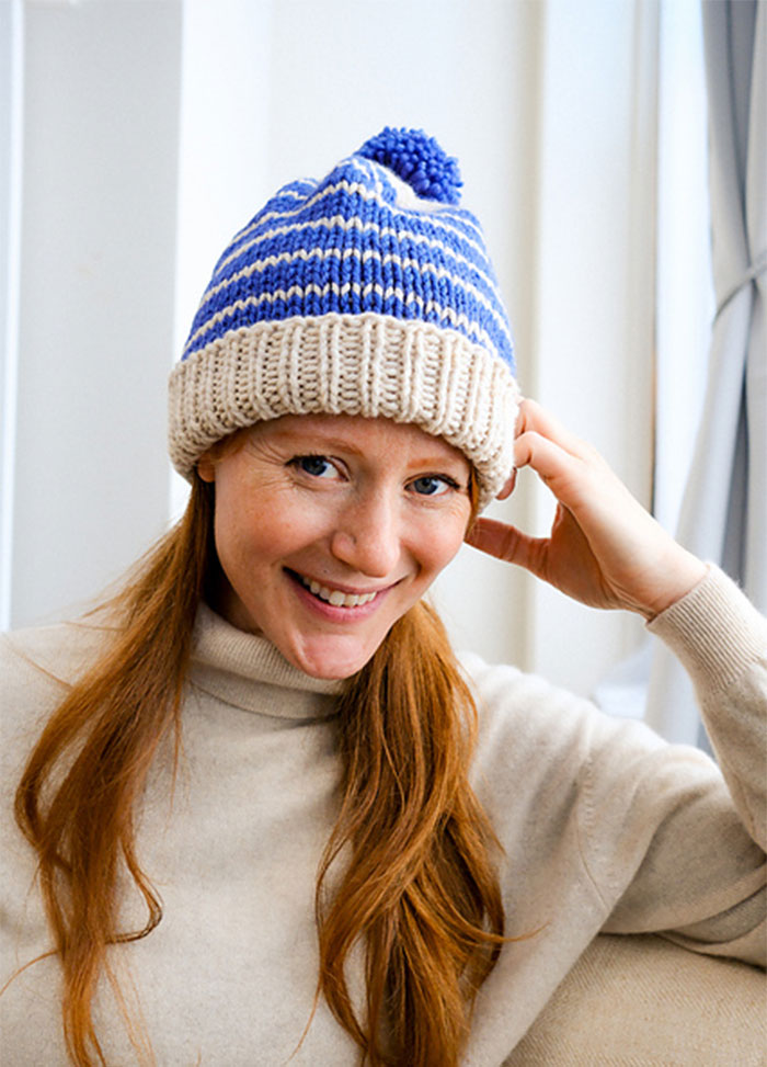 Model wearing a blue and white striped Cardo Holiday Hat with a blue pom-pom and beige ribbed brim, indoors with a light turtleneck sweater.