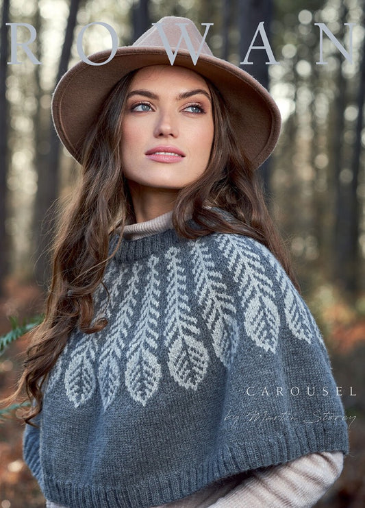 A woman in a tan hat and a cozy gray patterned sweater stands in a forest. Sunlight filters through tall trees, creating a serene, autumnal backdrop.