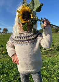 A child in a knitted sweater stands in a sunny field, holding a large sunflower in front of their face. The sky is clear blue, creating a cheerful atmosphere.