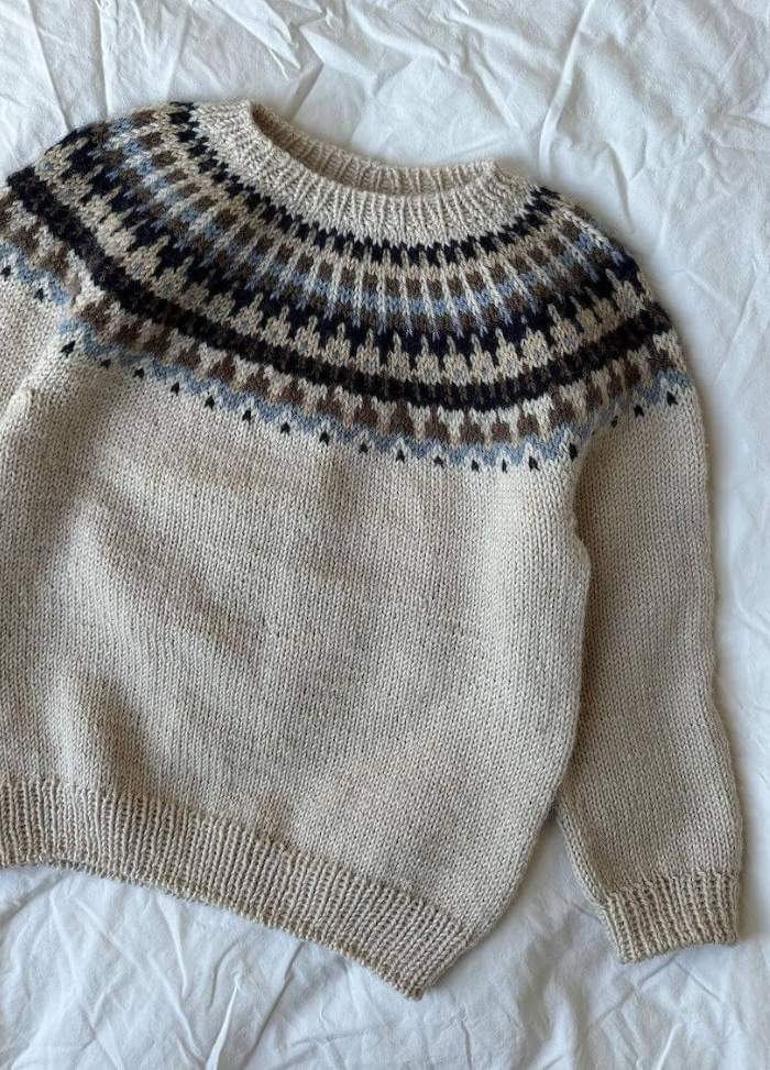 Beige knit sweater with a patterned yoke featuring brown, grey, and black geometric designs. The sweater sits on a crumpled white fabric backdrop.