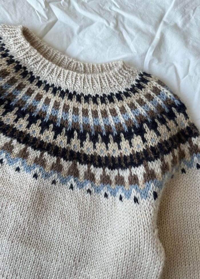 Close-up of a knitted sweater with an intricate, multi-coloured pattern at the yoke in brown, blue, and cream tones, laid on a white fabric background.