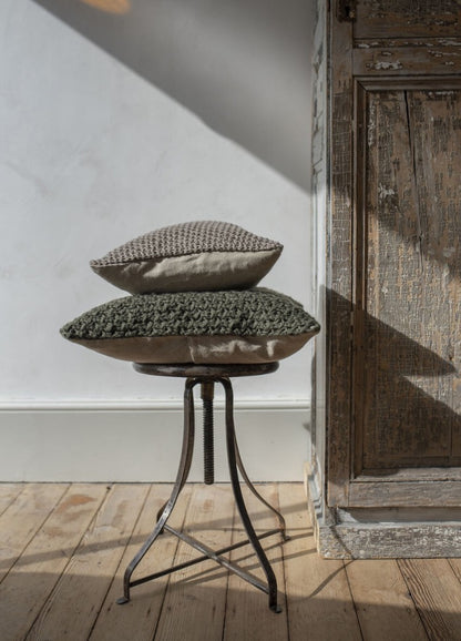 Stacked green and beige textured pillows rest on a rustic metal stool beside a weathered wooden door. Sunlight casts gentle shadows on the wooden floor.
