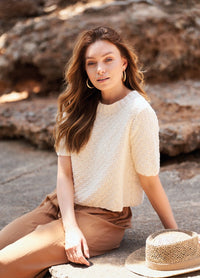 Woman with long hair sits outdoors on stone surface, wearing cream knit top and tan pants. She looks serene. Straw hat rests beside her. Sunny day.