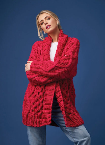 A woman stands confidently against a blue background, wearing a chunky red knit cardigan over a white top and jeans. Her arms are crossed and she has a poised expression.
