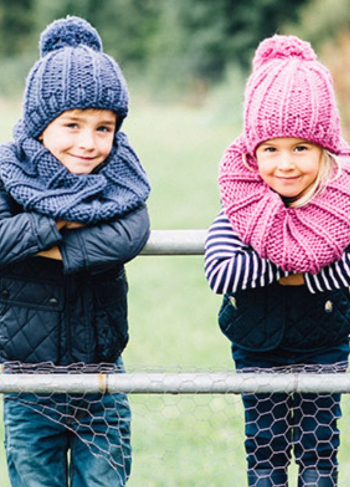 Charlie Cowl: Two children outdoors leaning on a metal fence, wearing hand-knitted cowls and hats‚Äîone in blue, the other in pink‚Äîwith a grassy landscape in the background.