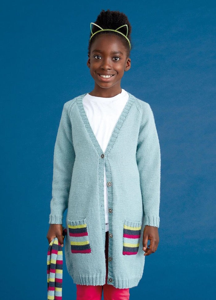 A child wearing a light blue cardigan with colorful pocket accents, red pants, and a cat-ear headband stands against a blue background, smiling warmly.