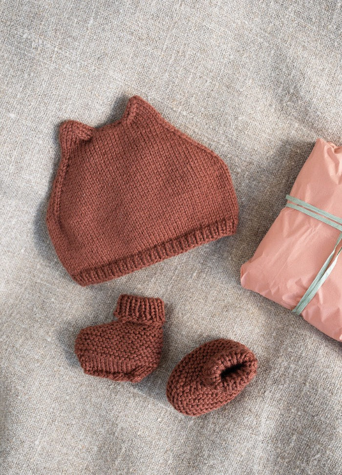 A knitted brown baby hat with ears and matching booties lie on tan fabric beside a pink, ribbon-tied package, evoking warmth and coziness.