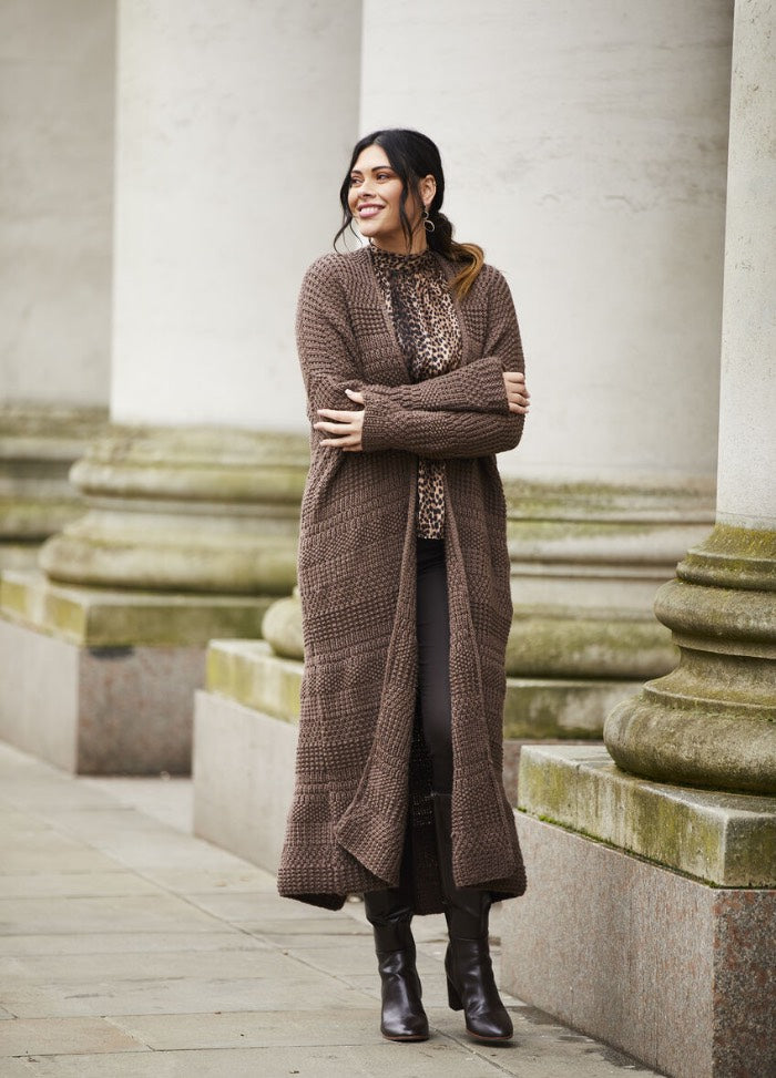 A smiling woman stands near large stone columns, wearing a long brown cardigan, leopard print top, and dark pants. The scene is elegant and serene.