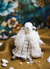 Agi the Sheep: knitted sheep in a checkered dress and fluffy shawl, posed on fabric with seashells and floral backdrop.