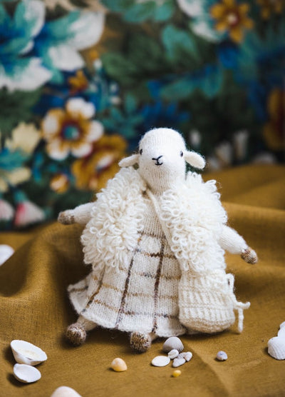 Agi the Sheep: knitted sheep in a checkered dress and fluffy shawl, posed on fabric with seashells and floral backdrop.