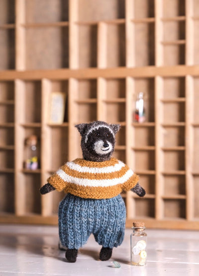 Billie the Raccoon: knitted raccoon in a striped sweater and blue pants, posed in front of a wooden shelf with jars and books.