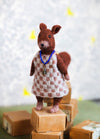 Hazel the Squirrel: knitted squirrel in a checkered dress and blue pendant, posed atop stacked cardboard boxes.