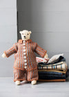 Mouche the Bear: knitted bear in a striped buttoned outfit, posed beside folded patterned fabrics.