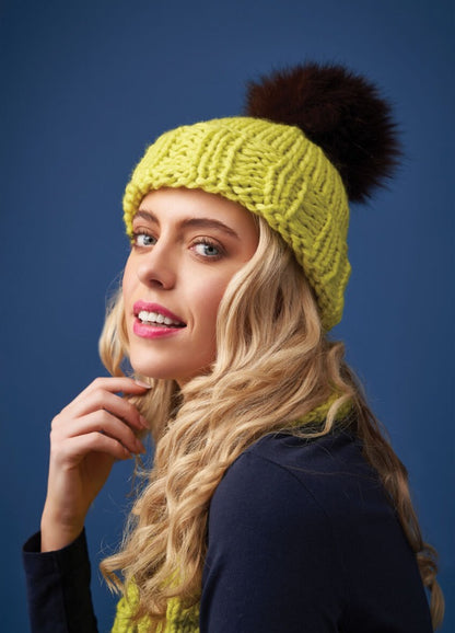 Blonde woman with curly hair wearing a bright yellow knit hat with a dark pom-pom, smiling against a blue background, conveying warmth and cheer.