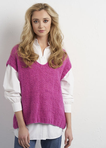 A woman with long, wavy blonde hair wears a bright pink, textured sweater over a white shirt and blue jeans, standing against a light background, exuding a casual yet stylish vibe.