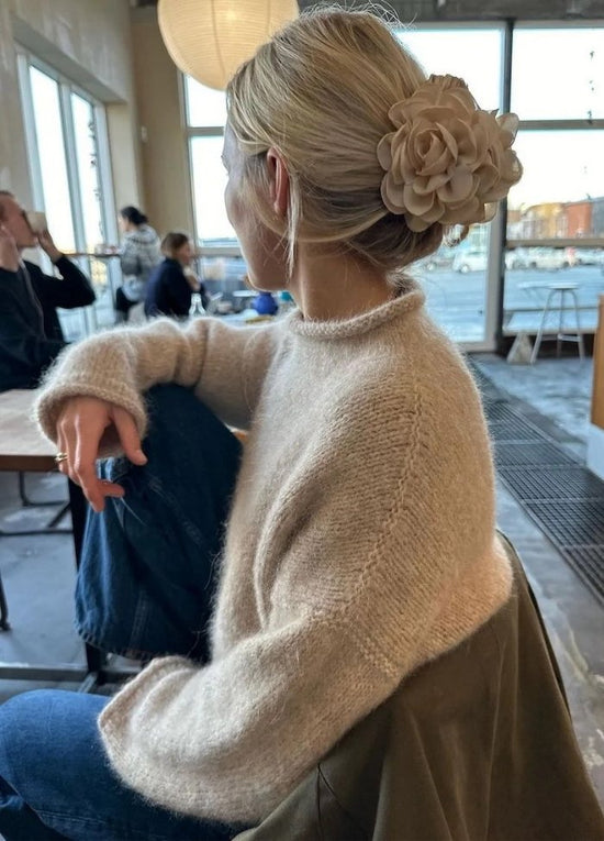 Woman in a cozy beige sweater with a large flower hair accessory sits in a cafe, looking outside. The atmosphere is warm and relaxed.