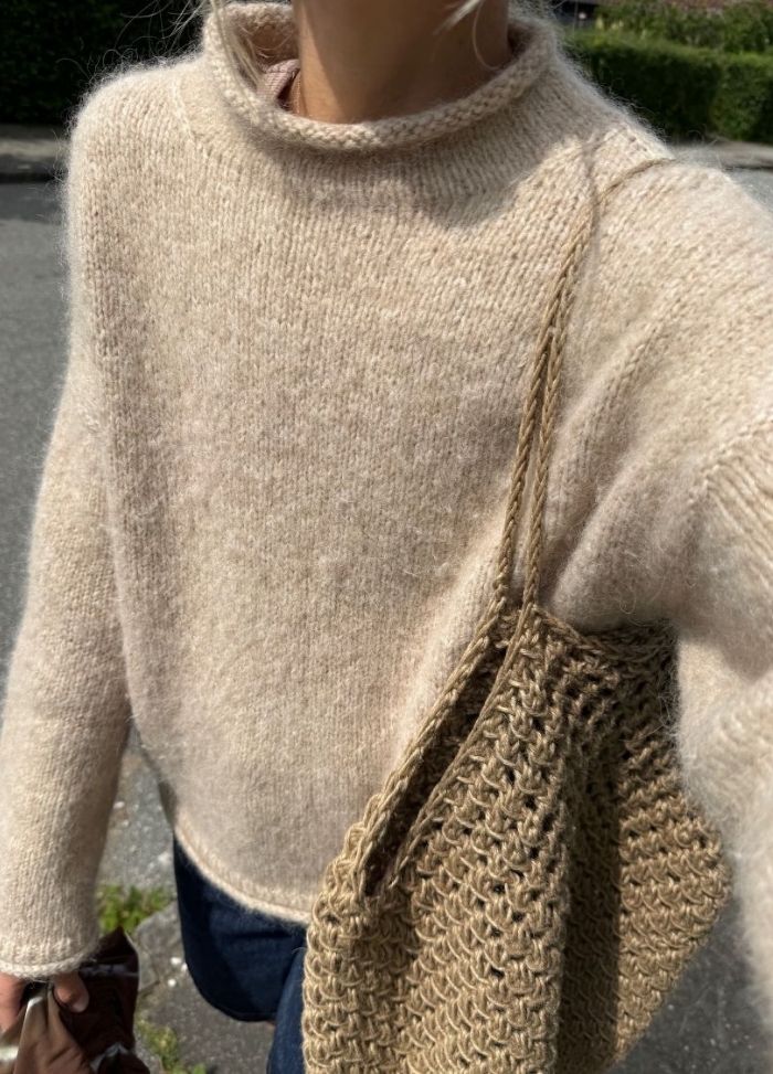 Person wearing a cozy, beige knit sweater with a high collar, carrying a textured straw bag. The scene has an outdoor, casual autumn vibe.
