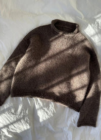 A cozy brown knit sweater lies on a textured white sheet, bathed in soft sunlight casting angled shadows.