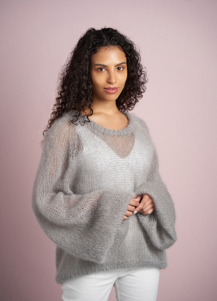 A woman with curly hair wears a cozy, loose-fitting gray sweater against a soft pink background, conveying warmth and confidence.