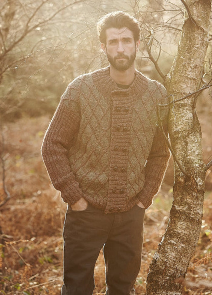 Person wearing a brown Clydesdale Cardigan with diamond pattern and ribbed sleeves, standing in a wooded outdoor setting.