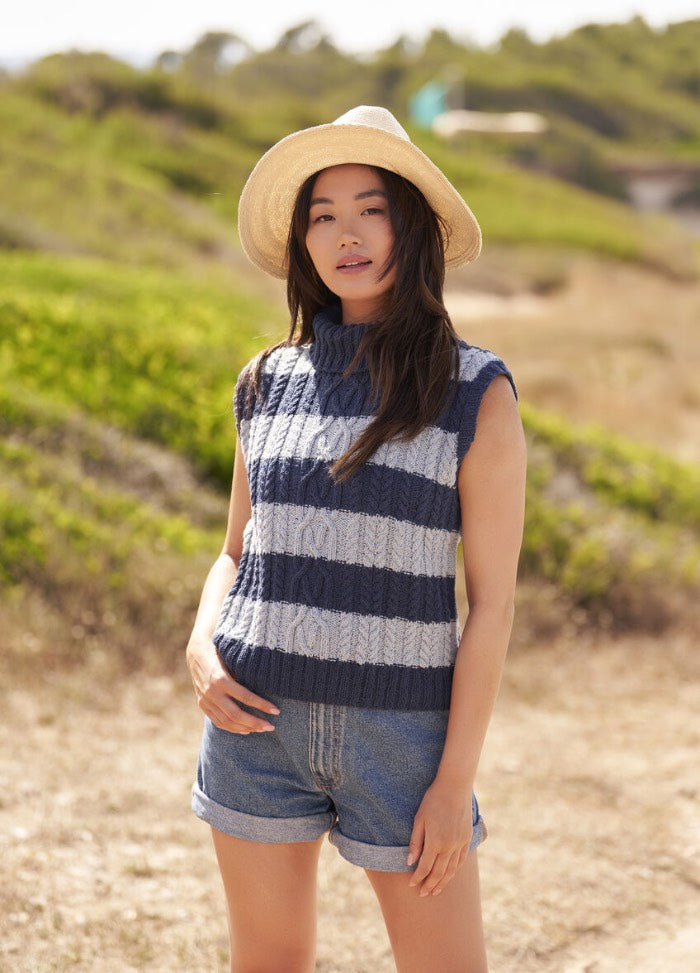 A woman in a wide-brimmed hat stands outdoors, wearing a sleeveless, striped knit sweater and denim shorts. The background is a sunlit, grassy landscape.