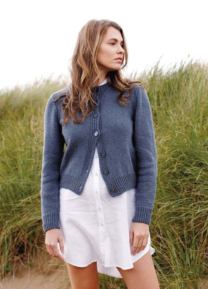 A woman stands in front of tall grass, wearing a blue cardigan and white dress. She looks to the side, with a contemplative expression, evoking a serene mood.
