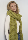 A woman with blonde hair leans against a white wall, wearing a cream top and a chunky green knit scarf. She looks serene, conveying a calm tone.