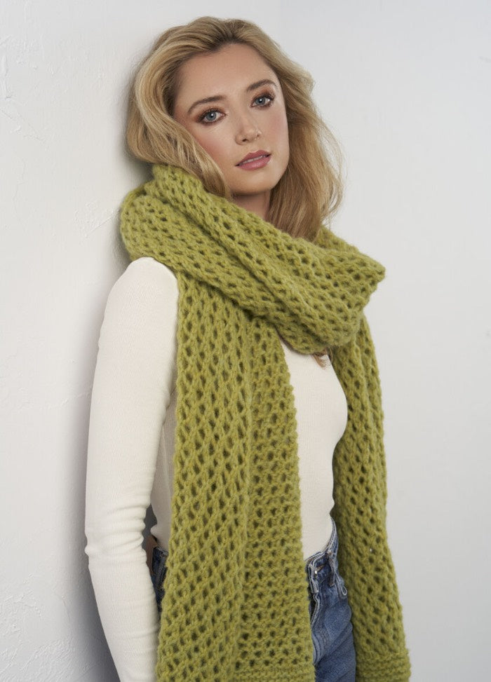 A woman with blonde hair leans against a white wall, wearing a cream top and a chunky green knit scarf. She looks serene, conveying a calm tone.