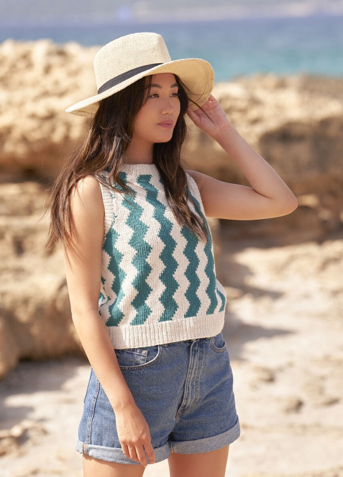 Woman in a straw hat and zigzag-patterned sleeveless top gazes to the side on a sunny beach. She wears denim shorts. Relaxed and serene.