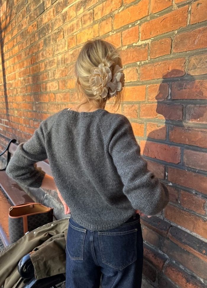 A person with blonde hair in a bun adorned with floral clips stands against a sunlit brick wall. They wear a grey sweater and blue jeans, conveying a casual, peaceful setting.