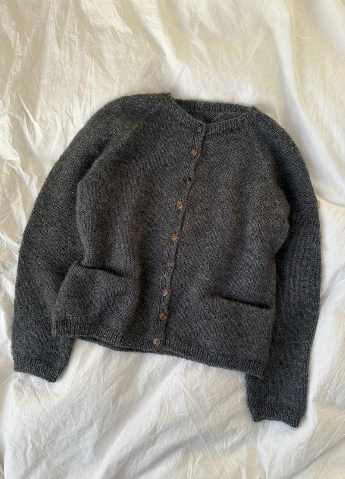 A dark grey knitted cardigan with brown buttons and pockets, laid on a white textured fabric. The cozy texture conveys warmth and comfort.