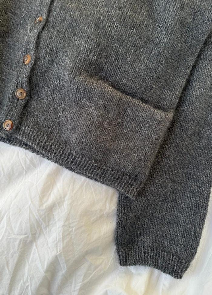 Close-up of a grey knitted cardigan with wooden buttons and a visible pocket on its side. It's laid out on a crinkled white fabric. Cozy and textured appearance.