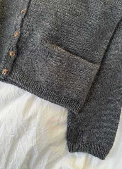 Close-up of a grey knitted cardigan with wooden buttons and a visible pocket on its side. It's laid out on a crinkled white fabric. Cozy and textured appearance.