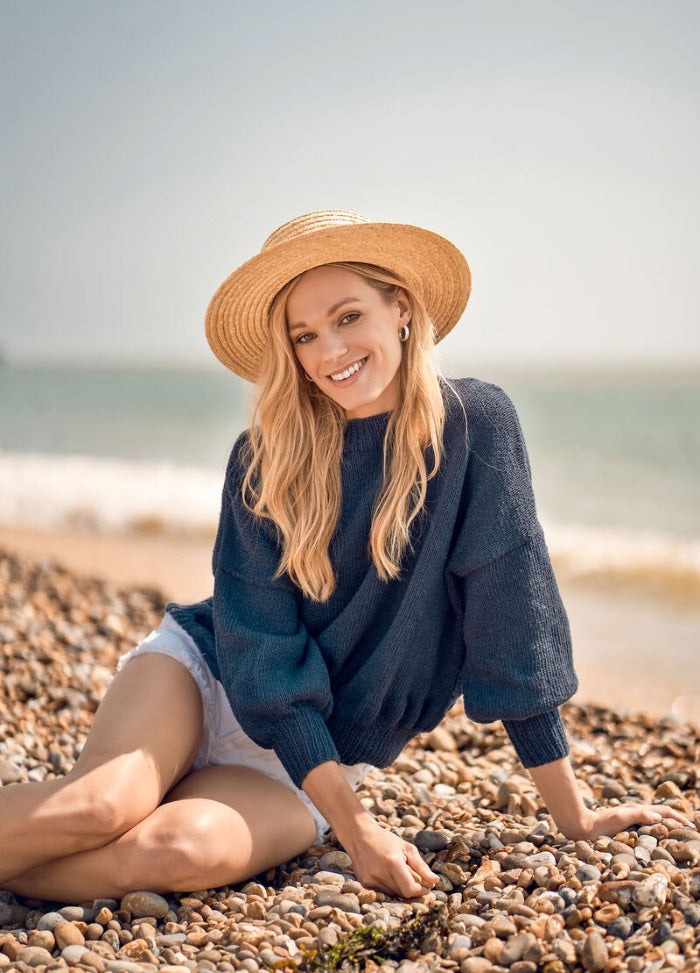 A woman in a straw hat and blue sweater smiles while sitting on a pebble beach. She wears white shorts, with waves gently breaking behind her.