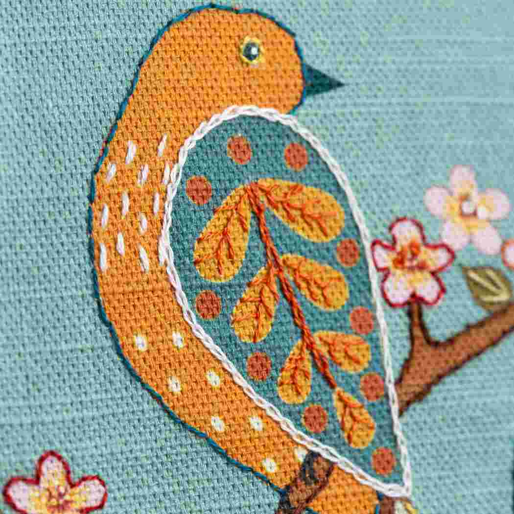 Cross-stitched orange bird on a branch with flowers on a light blue background