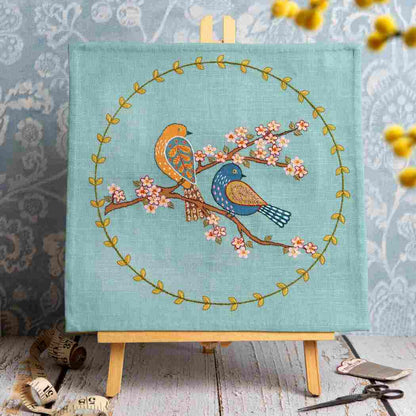 Embroidery of two birds on a branch with flowers on a teal background, displayed on a wooden easel.