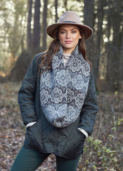 Woman in a quilted jacket and wide-brim hat stands in a forest. She wears a patterned scarf, exuding a calm and stylish autumn vibe.