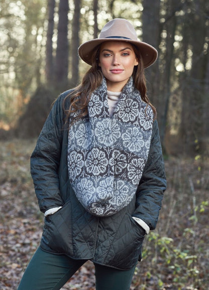 Woman in a quilted jacket and wide-brim hat stands in a forest. She wears a patterned scarf, exuding a calm and stylish autumn vibe.