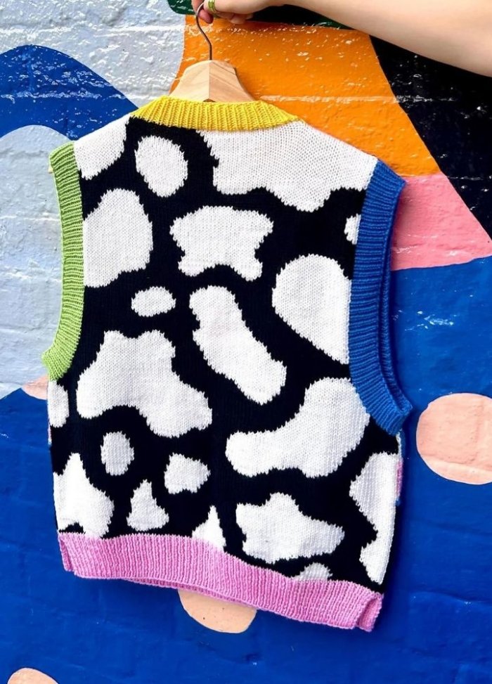 Knitted vest with black and white abstract pattern, vibrant trim in green, yellow, pink, and blue, against a colorful painted wall background.