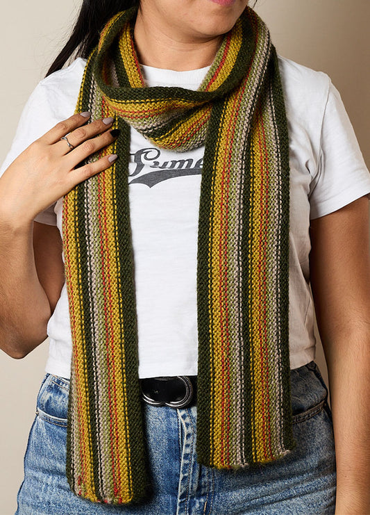 Person wearing a hand-knitted scarf with vertical stripes in green, yellow, orange, and gray, styled with a white t-shirt and blue jeans – vibrant and cozy accessory for everyday wear.