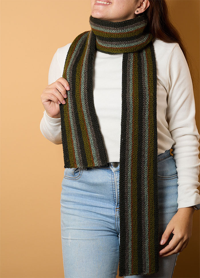 Person wearing a hand-knitted striped scarf in black, green, brown, and gray tones, paired with a white long-sleeve shirt and light blue jeans – cozy and colorful accessory against a light brown background.