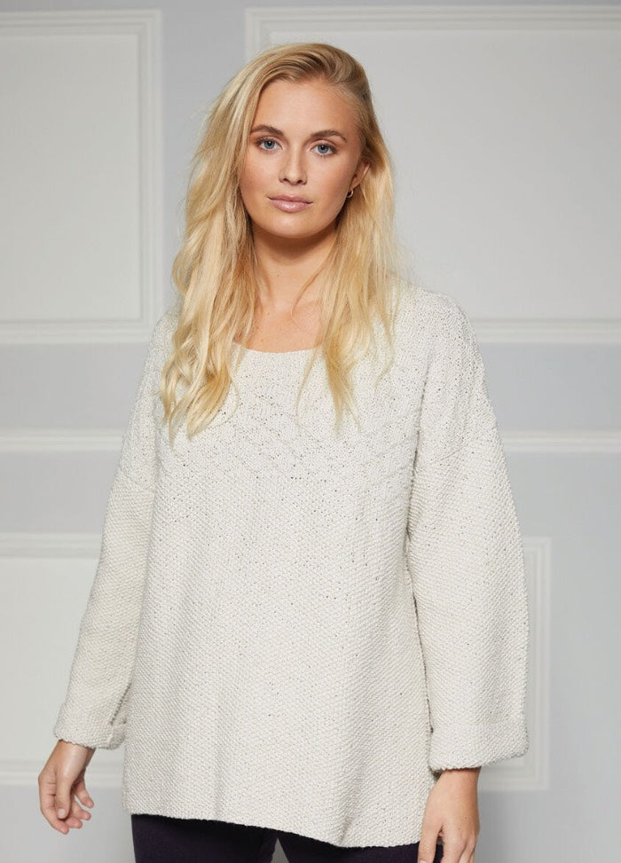 A person with long blonde hair wearing a cozy, light gray knit sweater stands against a light gray paneled wall, exuding a calm, relaxed demeanor.