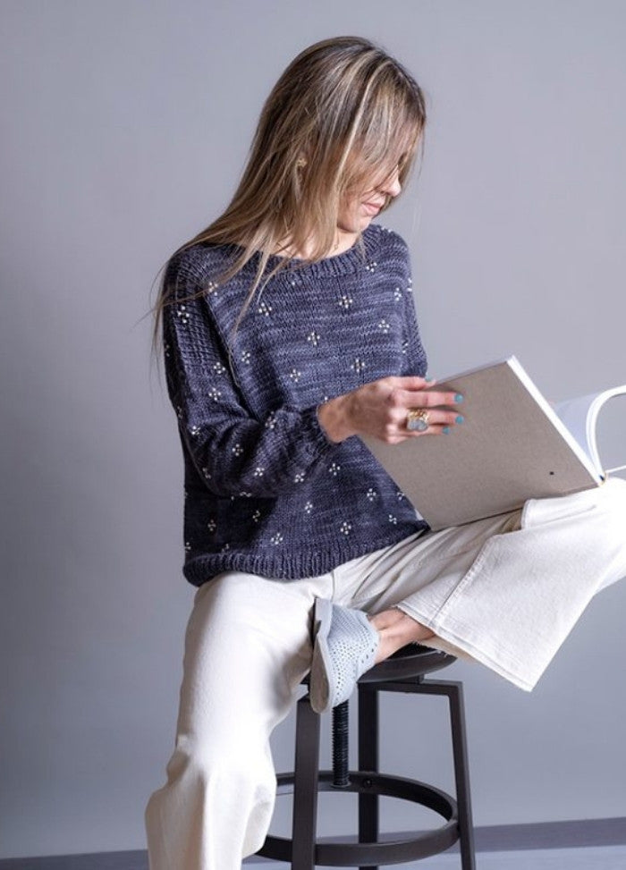 Woman with long hair sits on a stool, wearing a dark sweater and light pants. She's smiling while reading a book against a neutral background.