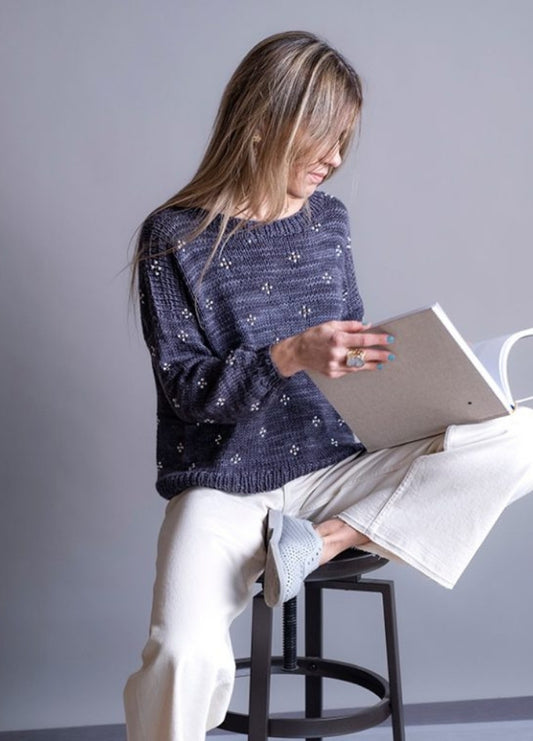 Woman with long hair sits on a stool, wearing a dark sweater and light pants. She's smiling while reading a book against a neutral background.