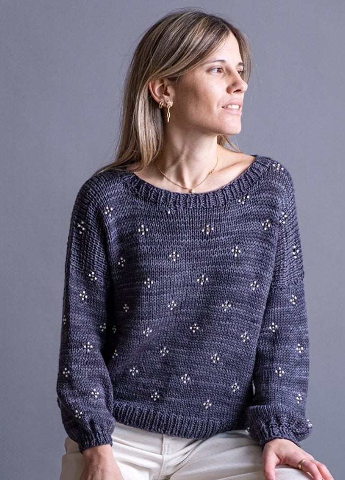 A woman in a gray, knitted sweater with subtle white patterns gazes to the side, smiling softly. She has light brown hair and wears gold earrings.
