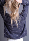 Person with long blonde hair pulled up, wearing a textured dark sweater with small white dots, standing against a gray background, facing away.