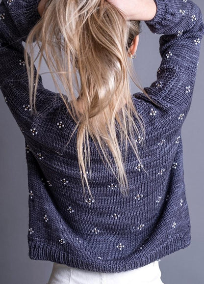 Person with long blonde hair pulled up, wearing a textured dark sweater with small white dots, standing against a gray background, facing away.