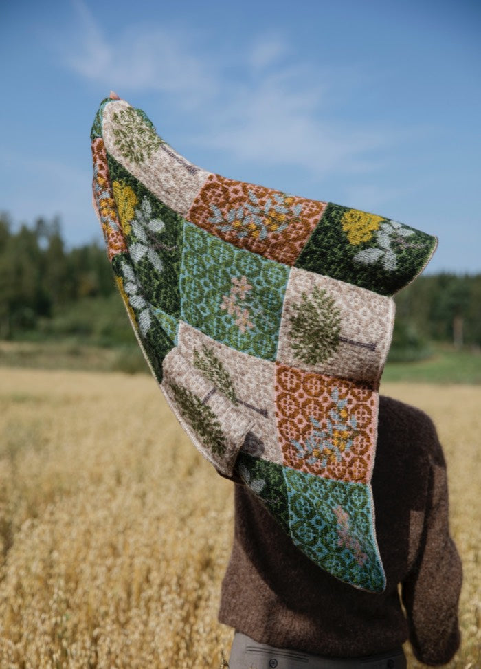 Parterre Scarf': Patchwork scarf with floral and geometric designs in green, brown, yellow, and blue, held up in a field.